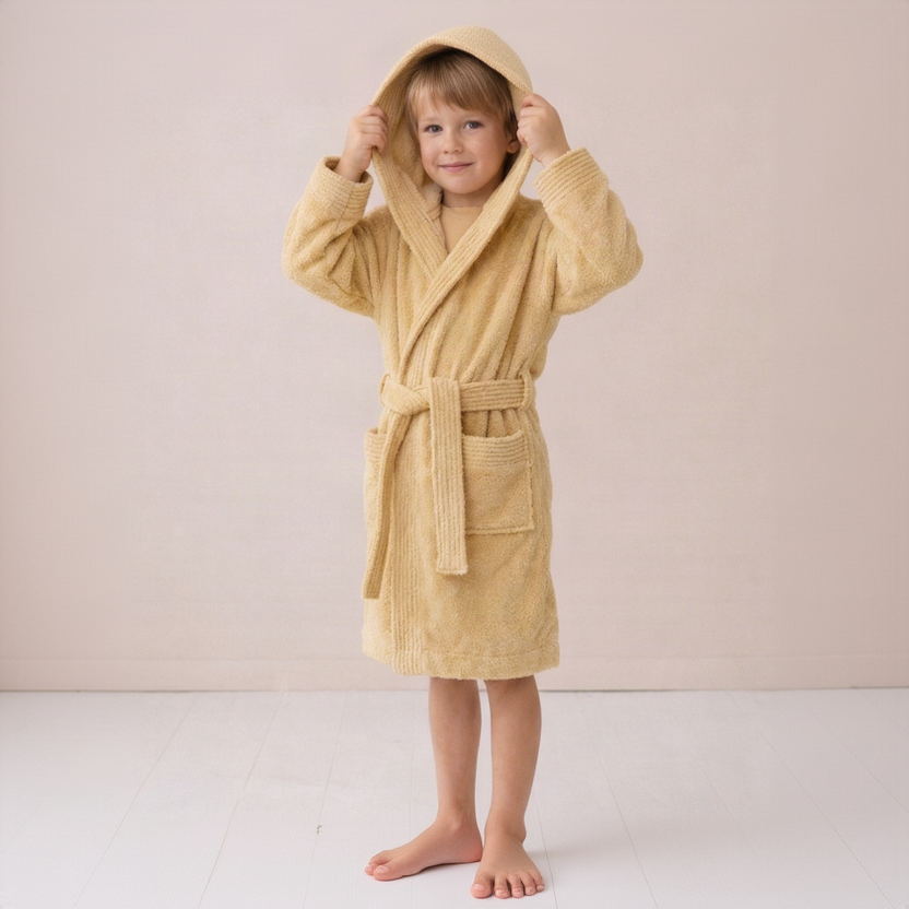A young child wearing a plush hooded bathrobe in a warm sandy beige color, standing barefoot against a soft, light beige studio background.