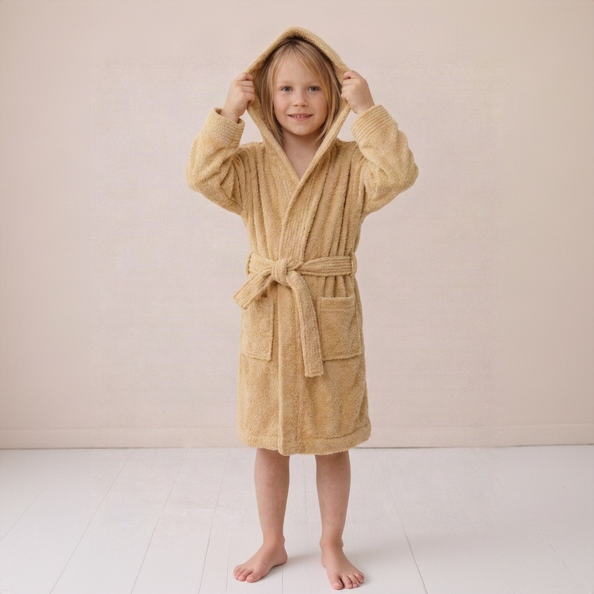A young child wearing a plush hooded bathrobe in a warm sandy beige color, standing barefoot against a soft cream-colored wall and light floor.