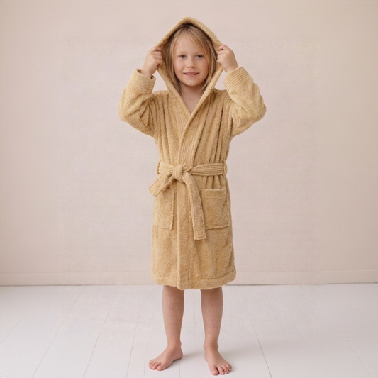 A young child wearing a plush hooded bathrobe in a warm sandy beige color, standing barefoot against a soft cream-colored wall and light floor.