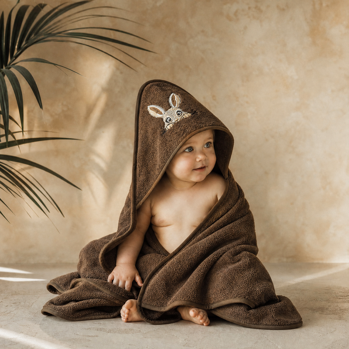 Child wrapped in a Dark Brown hooded towel with a deer design, sitting on a beige floor with a plant in the background.