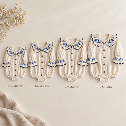 Four cream-colored baby long-sleeve bodysuits in different sizes arranged in a row on a light beige background, each with blue floral embroidery on the collar and cuffs, black scalloped trim, and front buttons. Text below each garment indicates the age range (0-3 Months, 3-6 Months, 6-9 Months, 9-12 Months).