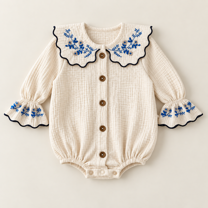 An ivory baby bodysuit made of soft muslin fabric, featuring a large scalloped Peter Pan collar with blue floral embroidery and navy trim, long sleeves with matching embroidered cuffs, and buttons down the front.