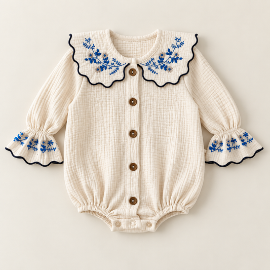 An ivory baby bodysuit made of soft muslin fabric, featuring a large scalloped Peter Pan collar with blue floral embroidery and navy trim, long sleeves with matching embroidered cuffs, and buttons down the front.