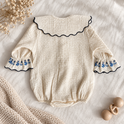 An ivory muslin baby bodysuit with a large scalloped Peter Pan collar trimmed in navy, and matching embroidered cuffs, laid flat on a white fabric background with baby props.