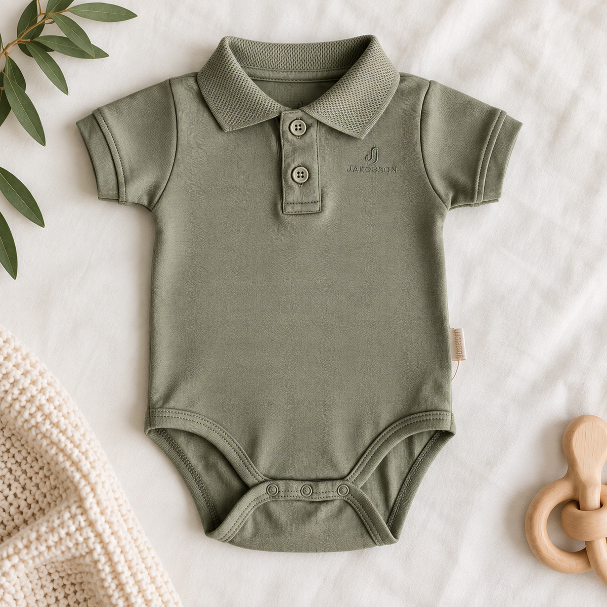 An olive green baby short-sleeve polo bodysuit with two buttons, laid flat on a white fabric background with decorative props including a knit blanket, green leaves, and a wooden pacifier.