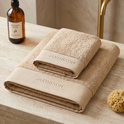 2-piece organic cotton towel set in beige with Jakobson branding displayed on a bathroom counter with a bottle and sponge
