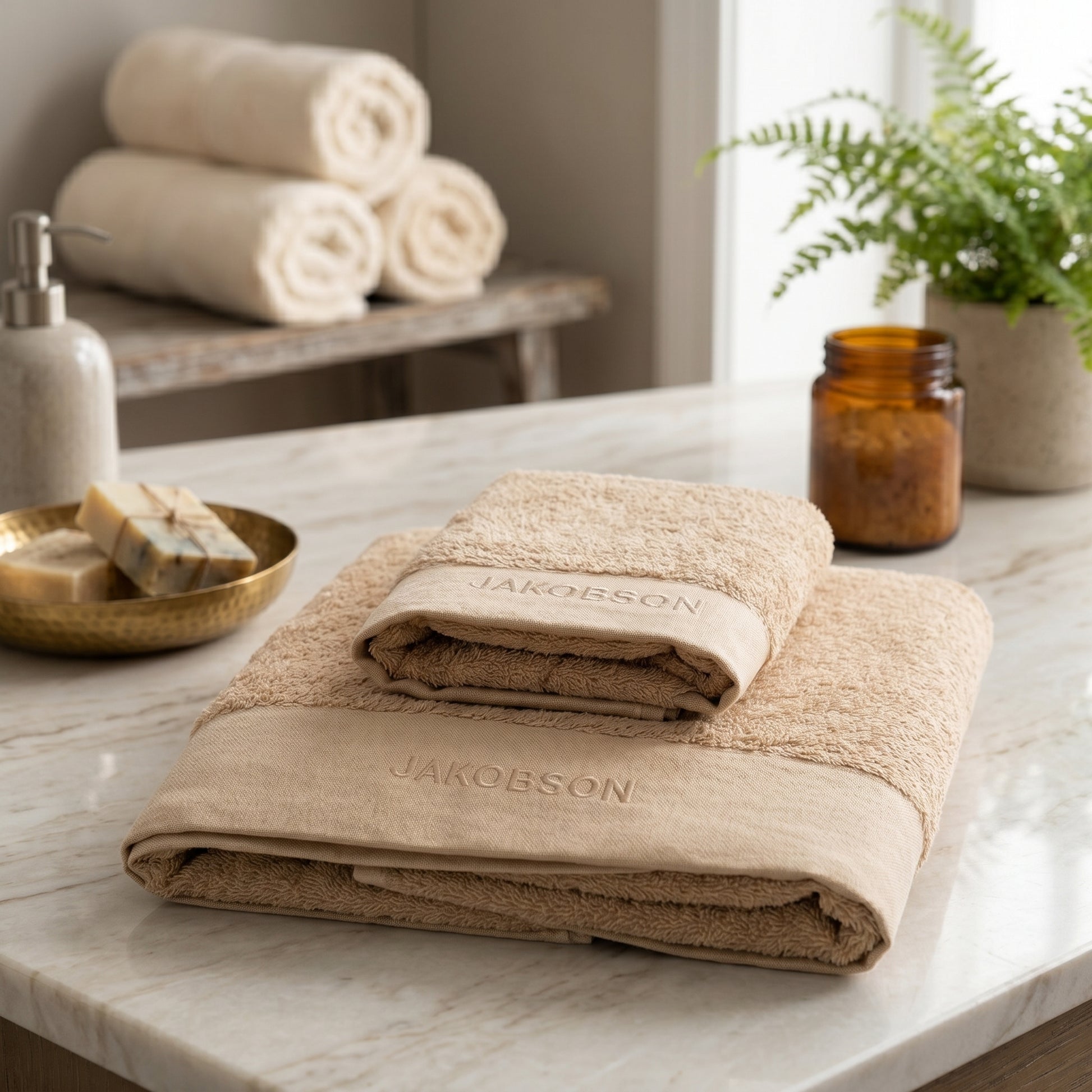 2-piece organic cotton towel set in beige with Jakobson branding displayed on a marble surface with a plant and soap dish