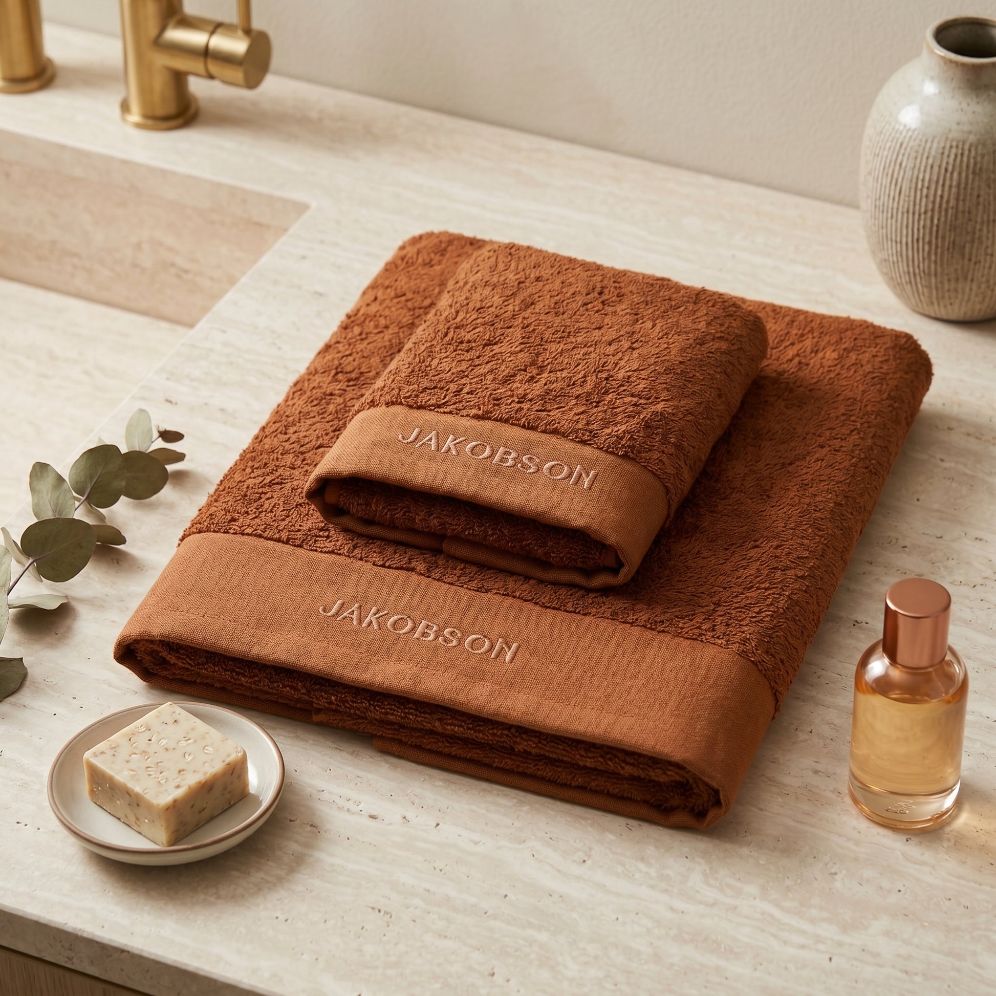 2-piece organic cotton towel set in cinnamon with Jakobson branding displayed on a bathroom counter with soap and bath accessories