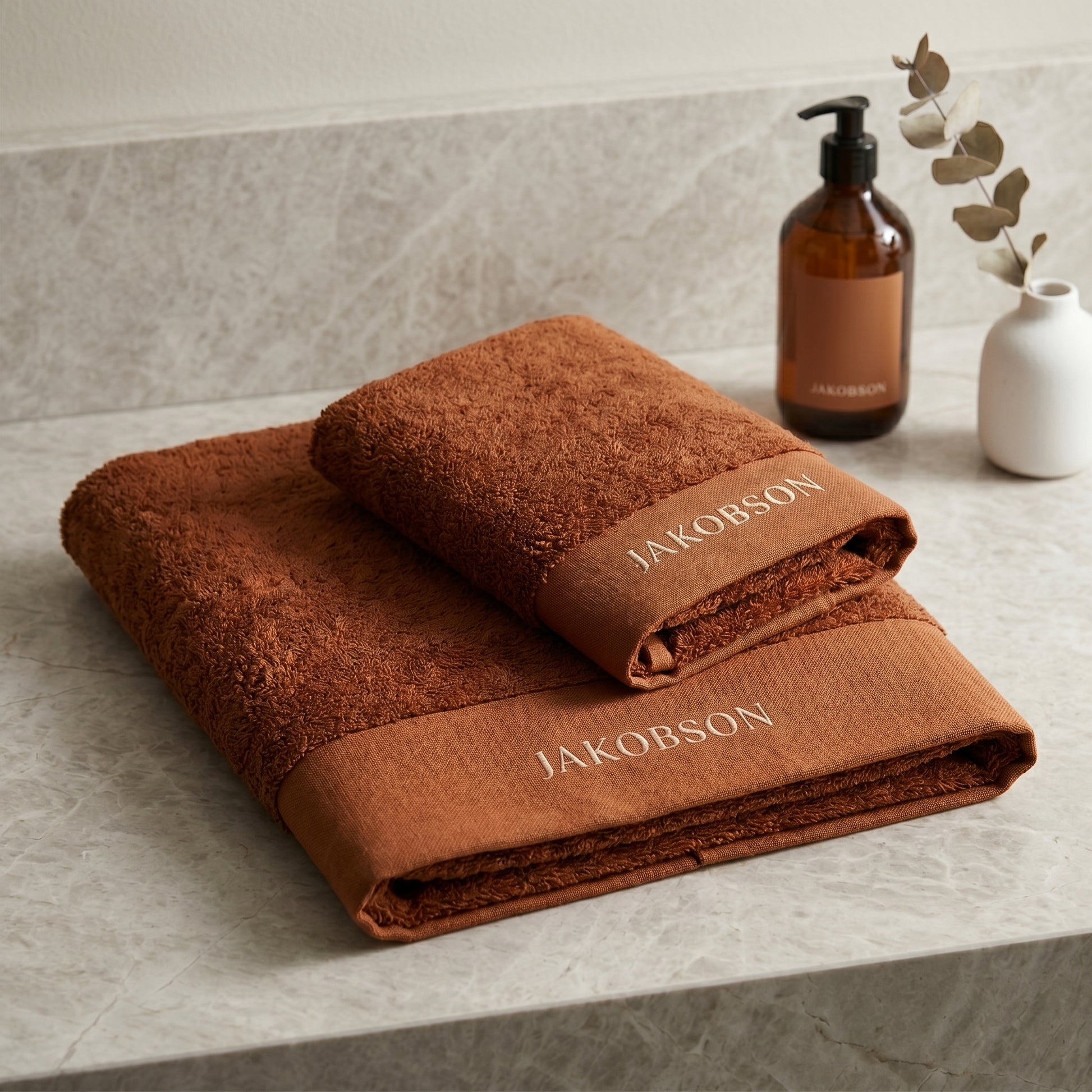 2-piece organic cotton towel set in cinnamon with Jakobson branding displayed on a marble surface with a bath bottle and decorative vase

