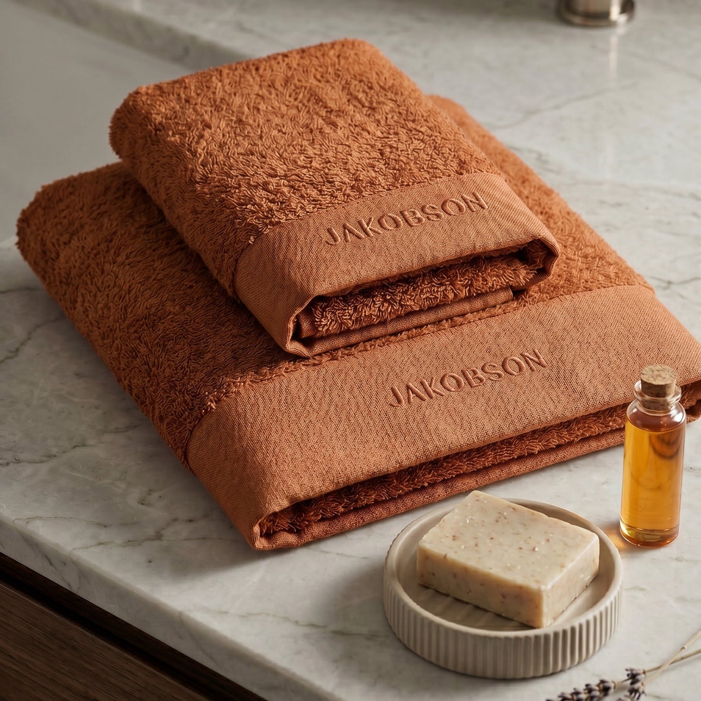 2-piece organic cotton towel set in cinnamon with Jakobson branding displayed on a marble surface with soap and bath accessories