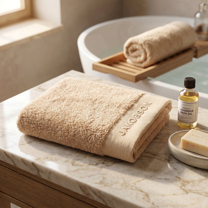 Organic cotton terry bath towel in beige with Jakobson branding displayed on a marble surface with lavender oil and soap