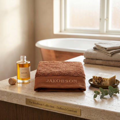 Organic cotton terry bath towel in cinnamon with Jakobson branding displayed with bath oil and soap