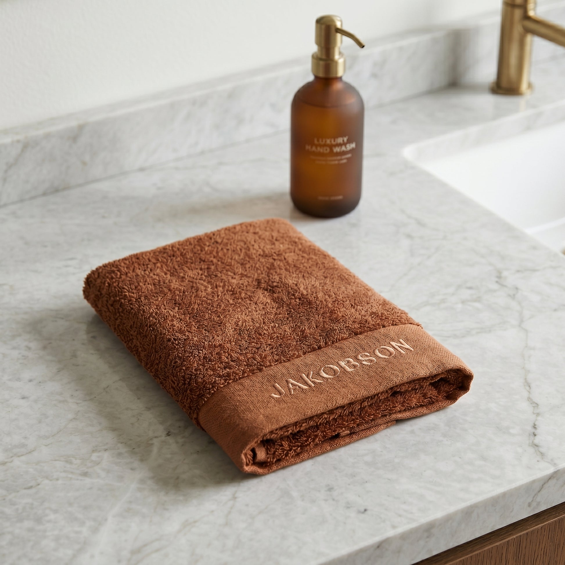 Organic cotton terry bath towel in cinnamon with Jakobson branding displayed on a marble countertop beside a bath bottle