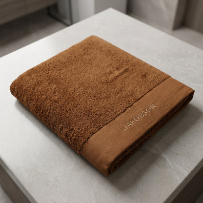 Organic cotton terry bath towel in cinnamon with Jakobson branding displayed on a marble surface