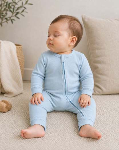 A baby wearing a light blue long-sleeve zip-front footless sleeper sits on a beige carpet in a cozy room setting with a pillow and blanket in the background.