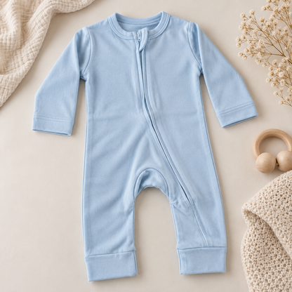 A baby long-sleeve footless zip-front romper in light blue, styled in a flat lay with a beige knit blanket, dried flowers, and a wooden ring toy on a warm beige background.