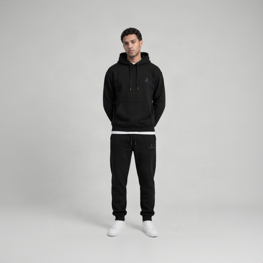 A man modeling a black pullover hoodie and matching black sweatpants with white sneakers, standing against a light gray studio background.