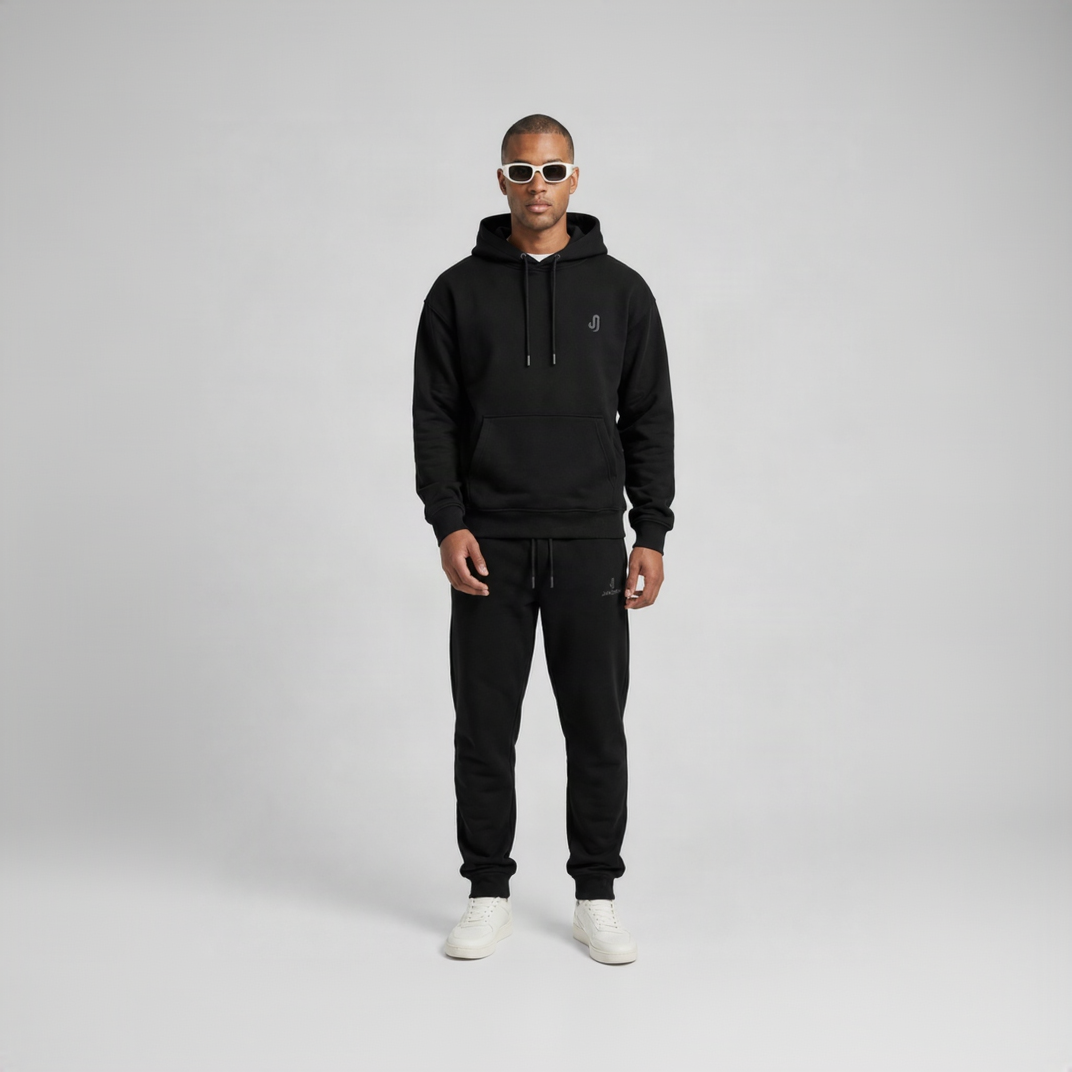 A man modeling a black pullover hoodie and matching black jogger sweatpants against a light gray studio background. The hoodie features a small white embroidered logo on the left chest, and the joggers have a small white logo on the thigh.