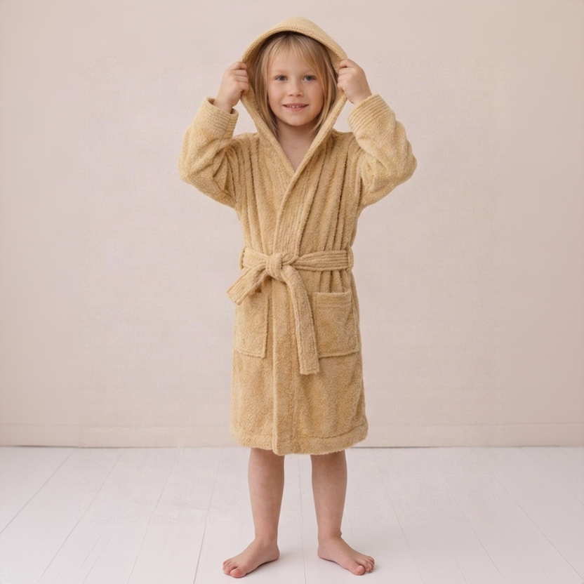 A young child wearing a plush hooded bathrobe in a warm sandy beige color, standing barefoot against a soft cream-colored wall and light floor.