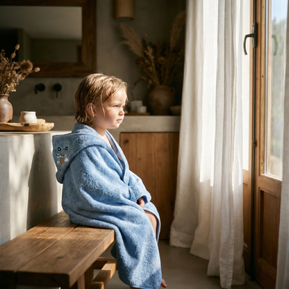 Child wearing a blue organic cotton hooded baby bath towel by a window